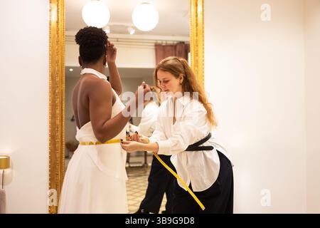 A tailor ensures a perfect fit by measuring a client waist in an elegant bridal fitting room. The scene reflects precision and attention to detail in Stock Photo