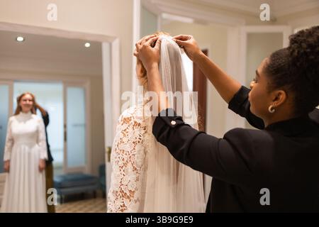 A bride stands in a bridal boutique mirror, admiring her lace gown, while a stylist adjusts her veil. The room elegant decor adds to the intimate wedd Stock Photo