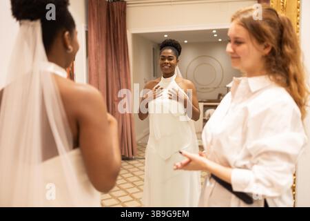 A bride in a fitting room joyfully admires her reflection in a white gown while a stylist looks on with a smile, creating a heartwarming, memorable mo Stock Photo