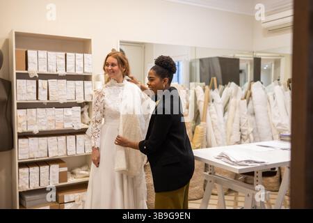In a bridal atelier, a bride is assisted by a stylist as she selects her wedding gown. Fabrics and lace details surround them, creating a cozy, elegan Stock Photo