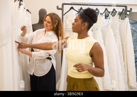 Two women engage in a thoughtful discussion while selecting elegant wedding dresses in a boutique. The setting exudes a sense of style, collaboration, Stock Photo