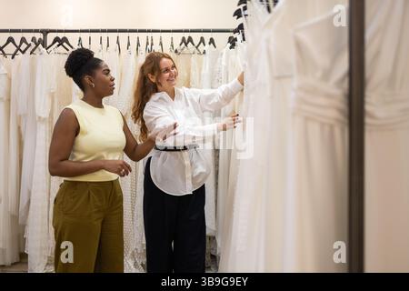 A bride and a consultant search through wedding dresses in a boutique. They are smiling and discussing the options, immersed in the shopping experienc Stock Photo