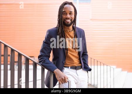 Cuban business man with long hair wearing a blazer and casual attire, leaning against a staircase railing outdoors, smiling warmly. Stylish urban sett Stock Photo