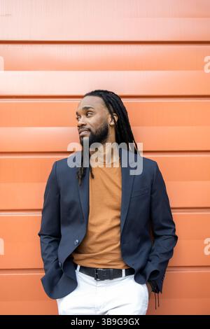 A cuban business man with dreadlocks leans against an orange wall, wearing a navy blazer and a brown shirt. He looks to the side, exuding confidence a Stock Photo