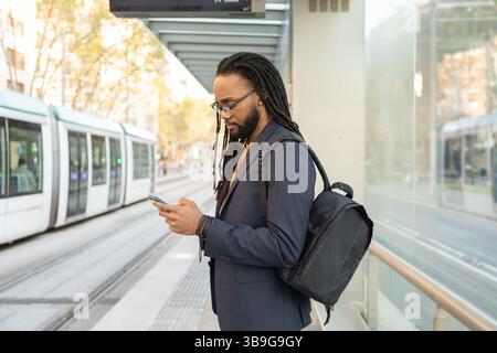 A cuban businessman in a suit, carrying a backpack, uses his phone while waiting at a tram stop. Captures the essence of modern urban life and technol Stock Photo