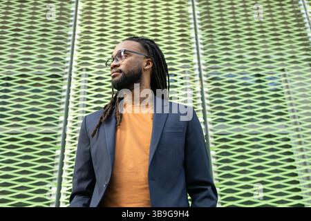 A cuban business man with long hair in glasses and a suit stands against a textured, patterned wall, embodying professionalism and style in an urban s Stock Photo