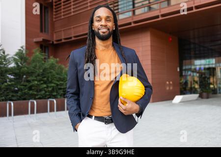A cuban businessman stands confidently outside a modern building, holding a yellow hard hat. He is dressed in a suit, representing expertise and profe Stock Photo
