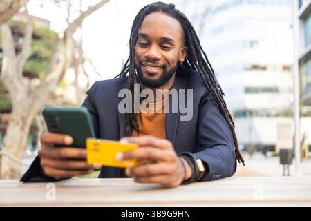 A cuban businessman with long hair smiles as he looks at his smartphone, holding a credit card. He is seated outdoors at a table, wearing a blazer. Br Stock Photo