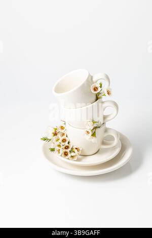 Stack of white ceramic cups and a saucer, elegantly garnished with fresh spring flowers on a white background, reflecting a serene and aesthetic vibe. Stock Photo