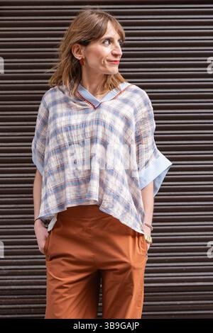 Stylish woman stands confidently in a plaid blouse and brown pants against a striped background. Her casual yet chic look exemplifies modern street we Stock Photo