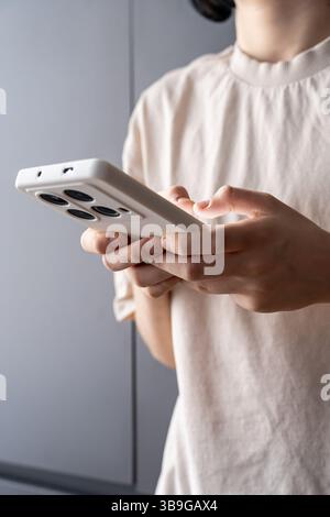 A teenager stands indoors using a smartphone with a white protective case, possibly typing or playing a game. The foreground highlights the interactio Stock Photo