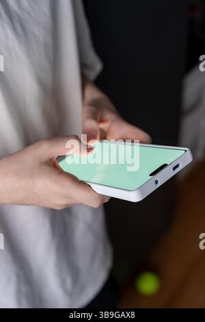 A teen stands indoors holding a smartphone with a bright screen, ideal for digital mockups or app interface design. The close-up highlights the user’s Stock Photo
