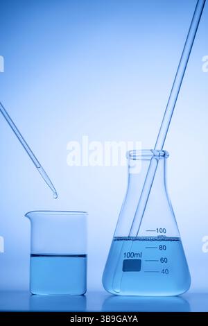 A laboratory beaker and flask filled with blue liquid, with a pipette dripping a drop in. The setup is illuminated with a soft blue light, giving a sc Stock Photo