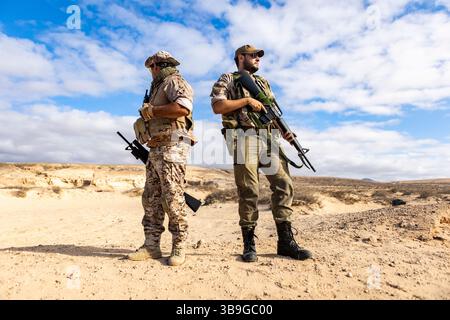 Two soldiers equipped with tactical gear and rifles stand back to back in a desert landscape, displaying teamwork and readiness under a clear blue sky Stock Photo