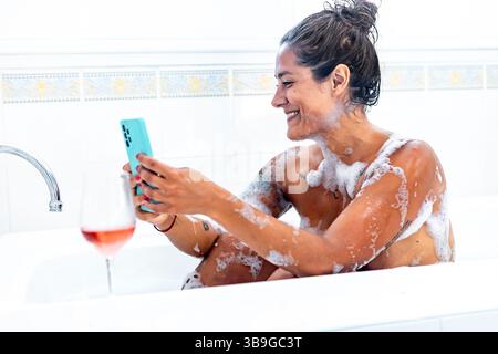 A woman relaxes in a bubble filled bath, using her smartphone with a smile. A glass of wine rests nearby, adding a touch of luxury to her serene momen Stock Photo