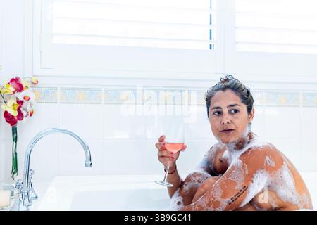 A woman enjoys a serene bubble bath, holding a glass of rose wine. Surrounded by suds and fresh flowers, she indulges in a moment of relaxation and se Stock Photo