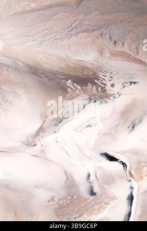 Aerial view capturing an abstract desert landscape. Soft sand dunes create wave patterns, blending tones of beige, cream, and light brown, invoking a Stock Photo