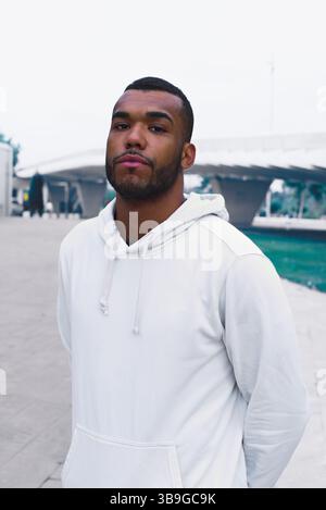 Portrait of a confident black male athlete wearing a white hoodie, posed outdoors in an urban environment. His strong presence and casual style convey Stock Photo