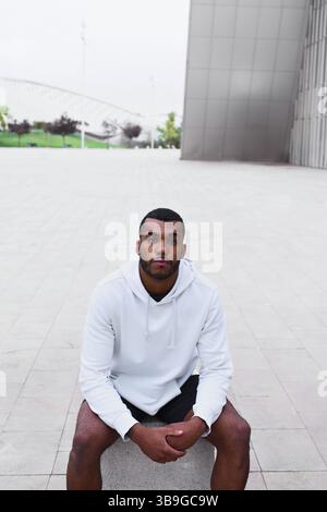 A young black male athlete sits outdoors in a modern urban environment, wearing a white hoodie and black shorts. The setting is minimalistic, echoing Stock Photo