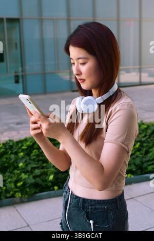 A young woman with headphones around her neck is focused on her smartphone. She stands outdoors with a modern building and greenery in the background. Stock Photo