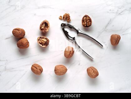 A scattered arrangement of whole and cracked walnuts with a metal nutcracker on a smooth marble surface, lit by natural light to highlight textures an Stock Photo