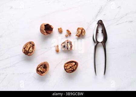 Cracked walnuts and a metal nutcracker on a white marble countertop. The arrangement highlights the rustic texture of the walnuts and the sleek design Stock Photo
