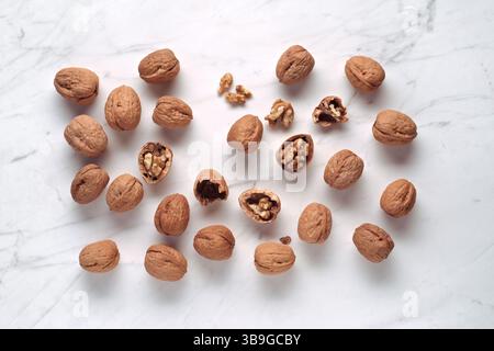 A collection of walnuts, some cracked open to reveal the kernels inside, scattered on a white marble surface. The nuts display their rich brown shells Stock Photo