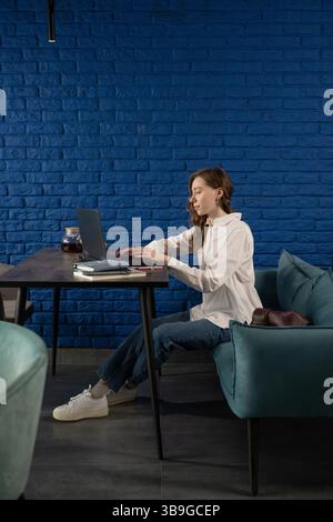 A woman sits in a modern blue cafe, working on a laptop at a black table. She is focused, wearing a white shirt and jeans, with notebooks and tea near Stock Photo