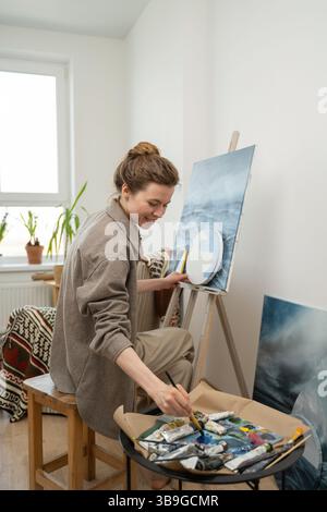 Artist happily working on a landscape painting in a bright, cozy home studio. Paints and brushes are seen on the table, emphasizing creativity and ins Stock Photo