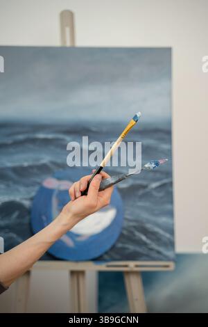 A close up of an artist hand holding a paintbrush and palette knife against a blurred ocean themed canvas. The scene captures the essence of creativit Stock Photo