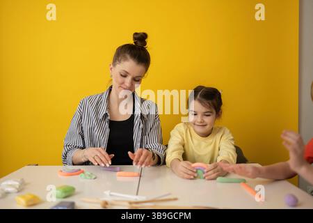 A woman and child create colorful clay shapes at a white table against a vibrant yellow background, encouraging creativity and learning through play i Stock Photo