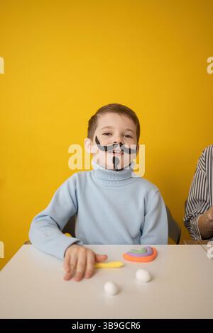 A boy with a playful clay mustache engages in creative activities at a white table, surrounded by colorful modeling clay, against a bright yellow back Stock Photo
