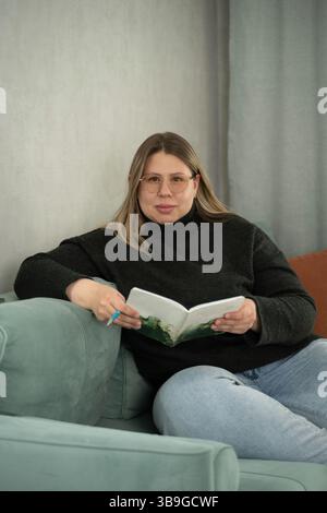 A woman in glasses relaxes on a soft sofa holding an open notebook and pen, dressed casually in a sweater and jeans. The calm setting and soft colors Stock Photo
