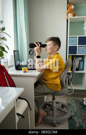 A curious boy in a yellow shirt sits barefoot at his desk using a monocular. Surrounded by a laptop, books, and toys, he explores and observes from th Stock Photo