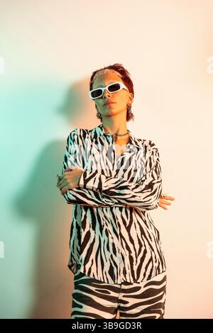 Young woman exuding confidence in bold zebra print attire, wearing trendy white sunglasses. She showcases a distinct modern style against a softly lit Stock Photo