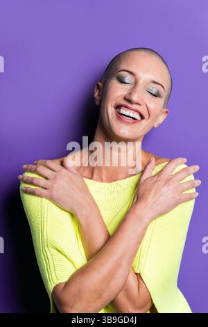 Middle aged shaved head woman in a vibrant yellow sweater, smiling broadly with eyes closed, exuding happiness and confidence against a bold purple ba Stock Photo