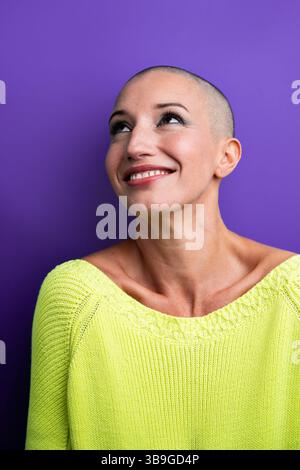 A joyful middle-aged woman with a shaved head smiles while looking upward, wearing a vibrant green sweater against a bold purple background, exuding c Stock Photo