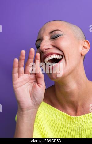 A joyful middle aged woman with a shaved head is captured in a moment of laughter, radiating happiness and confidence against a vibrant purple backdro Stock Photo