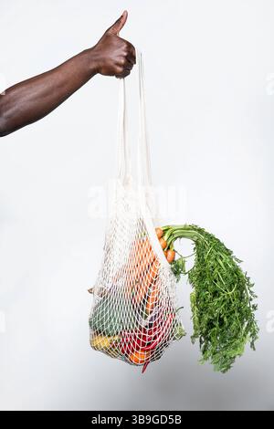 A person gives a thumbs-up while holding a mesh bag filled with fresh vegetables, symbolizing eco-friendly shopping and healthy living on a light back Stock Photo