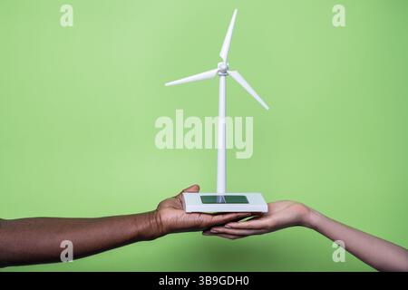 Close-up of diverse hands holding a small wind turbine model against a vibrant green background, symbolizing unity and commitment to sustainable and r Stock Photo
