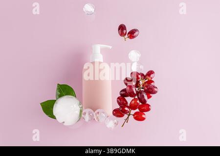 A stylish shower gel bottle surrounded by fresh red grapes, ice cubes, and green leaves on a pastel pink background, conveying freshness and elegance Stock Photo