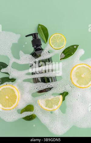 A black bottle of shower gel surrounded by vibrant lemon slices, fresh green leaves, and white foam on a mint green background, creating a refreshing Stock Photo
