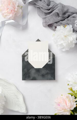 A chic wedding stationery arrangement featuring a blank card in a dark envelope, surrounded by delicate pink and white flowers and a soft gray ribbon Stock Photo