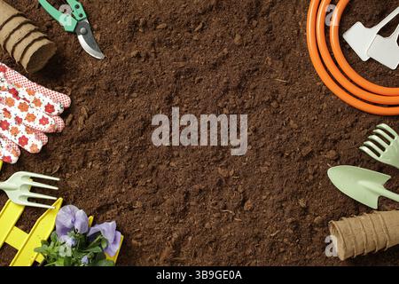 Various gardening tools and essentials, including gloves, a hose, and shovels, arranged on rich, brown soil, ready for planting flowers and maintainin Stock Photo