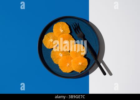 A minimalist setup showcasing vibrant orange citrus slices on a blue plate with a black fork, set against a striking blue and white background, creati Stock Photo