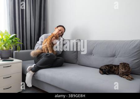 A relaxed woman cuddles her ginger cat on a gray couch, while another cat lounges beside her in a cozy home setting with neutral decor and a touch of Stock Photo