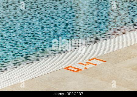 The image shows a swimming pool with clear blue water, bordered by a distinct checkered mosaic tile pattern. The edge features safety tiles and bright Stock Photo