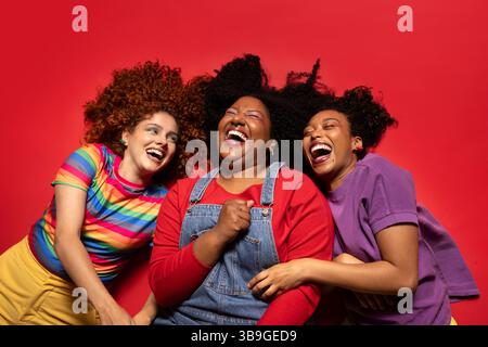 Three friends share a moment of pure joy and laughter, lying on a vibrant red background. Their colorful clothing and genuine expressions highlight th Stock Photo