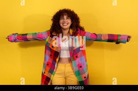 A cheerful woman stands with open arms, donning a vibrant, multicolored coat against a bright yellow background, radiating happiness and a lively spir Stock Photo