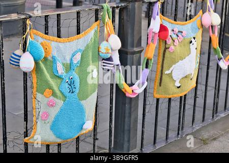 Knitted Easter decorations hang from a black metal fence. Two panels show a rabbit and a llama, with additional knitted eggs and a chain in Ripon, Yor Stock Photo